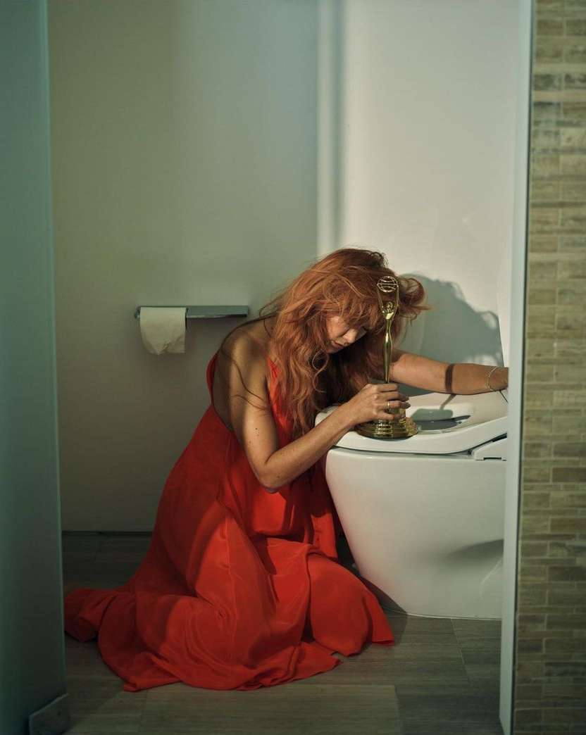 Woman in red dress kneeling in a bathroom, holding golden trophy, looking down with soft, diffused light and neutral color palette.