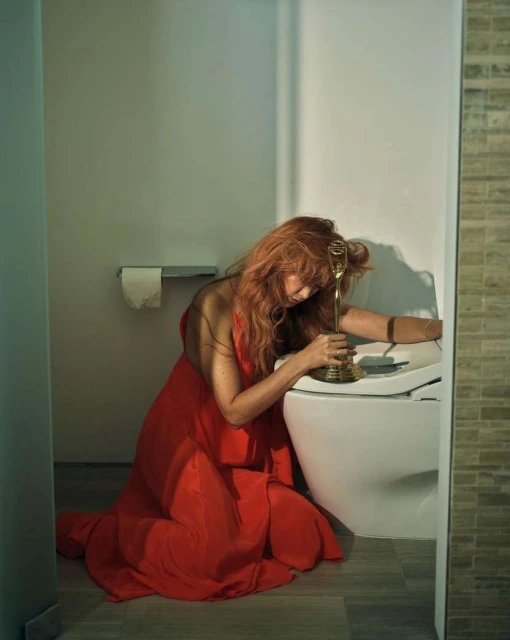 Woman in red dress kneeling in a bathroom, holding golden trophy, looking down with soft, diffused light and neutral color palette.