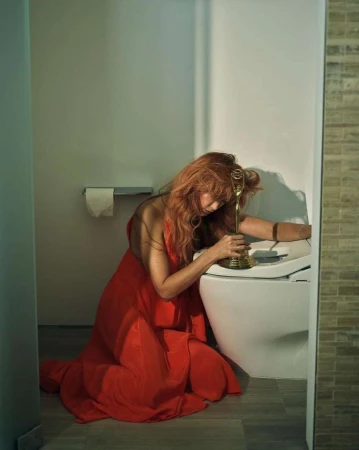 Woman in red dress kneeling in a bathroom, holding golden trophy, looking down with soft, diffused light and neutral color palette.