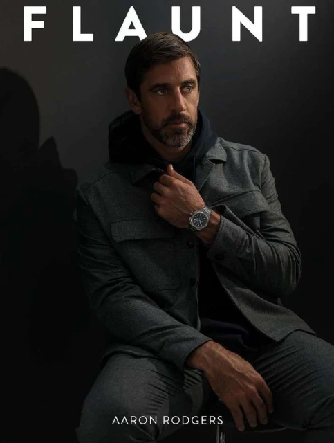 Aaron Rodgers poses in a gray jacket and black hoodie in a dimly lit photo studio setting for a magazine cover, looking off-camera.