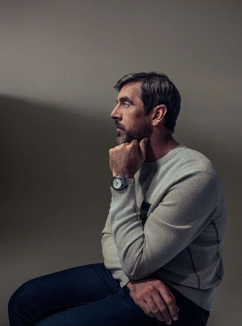 Profile portrait of a thoughtful man seated, wearing a sweater and watch, with a beard and graying hair against a neutral background.
