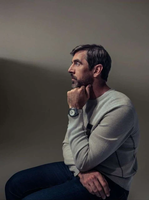 Profile portrait of a thoughtful man seated, wearing a sweater and watch, with a beard and graying hair against a neutral background.