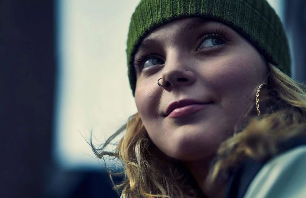 Close-up portrait of an adult female with blond hair wearing a green beanie, nose ring, and gold earrings, looking up and to the right.