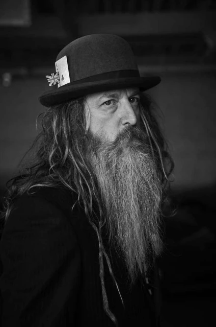 Portrait of a bearded man with long hair wearing a hat with a British flag, dressed in black, in a black and white photography.