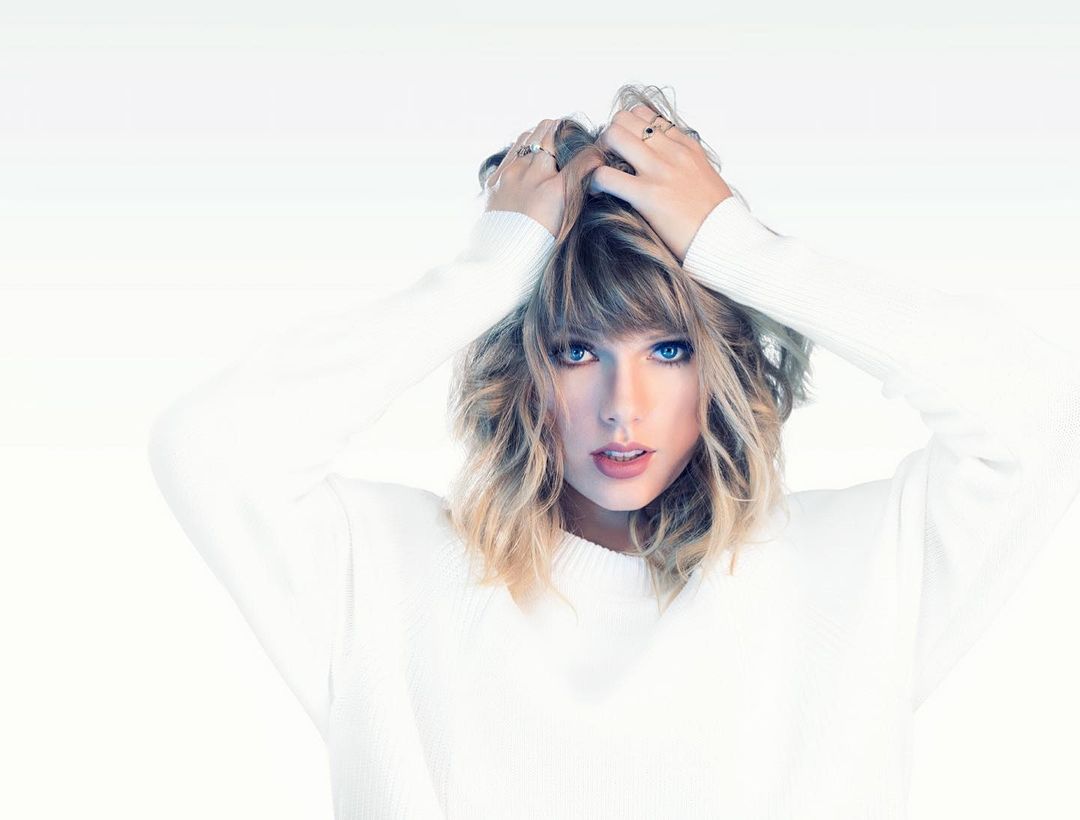 Blonde woman with wavy hair poses in a white sweater against a white background, holding her hands on top of her head.