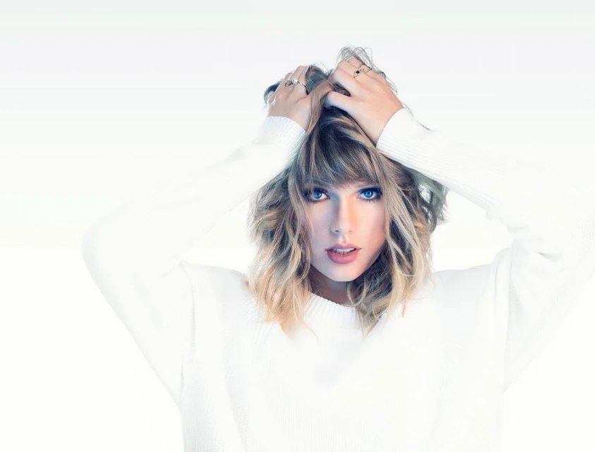 Blonde woman with wavy hair poses in a white sweater against a white background, holding her hands on top of her head.
