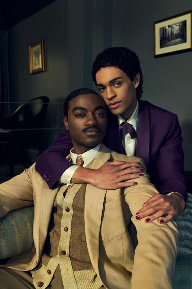 Portrait of two stylish adult males embracing indoors. One man wears a tan suit, while the other wears a purple suit with matching tie.