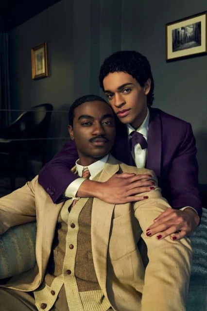 Portrait of two stylish adult males embracing indoors. One man wears a tan suit, while the other wears a purple suit with matching tie.