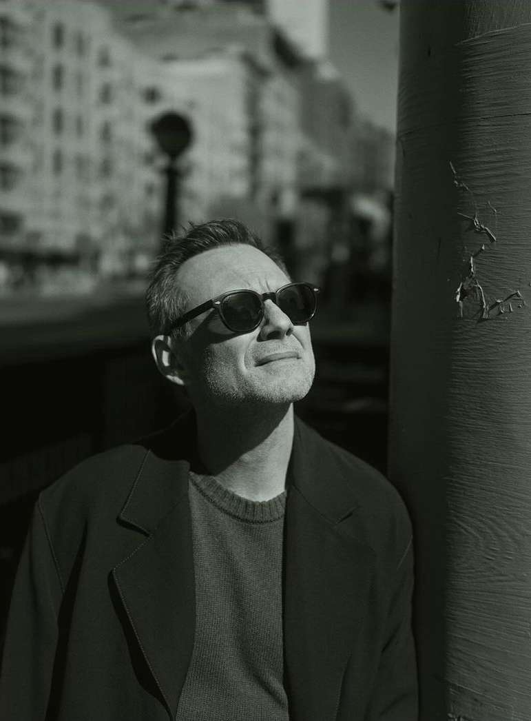 Monochrome portrait of a man in sunglasses and dark coat leaning against a pillar on a city street.