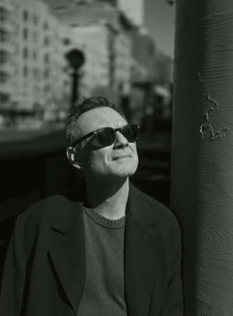 Monochrome portrait of a man in sunglasses and dark coat leaning against a pillar on a city street.
