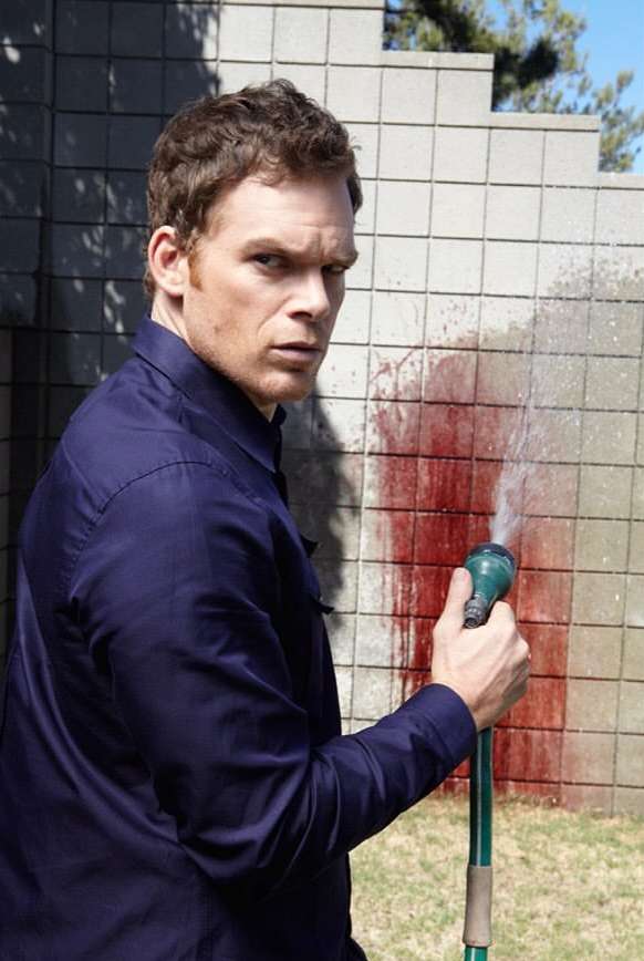 Man in a blue shirt outdoors with a garden hose pointed at red liquid resembling blood on a grey concrete wall.
