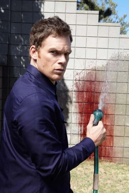 Man in a blue shirt outdoors with a garden hose pointed at red liquid resembling blood on a grey concrete wall. 