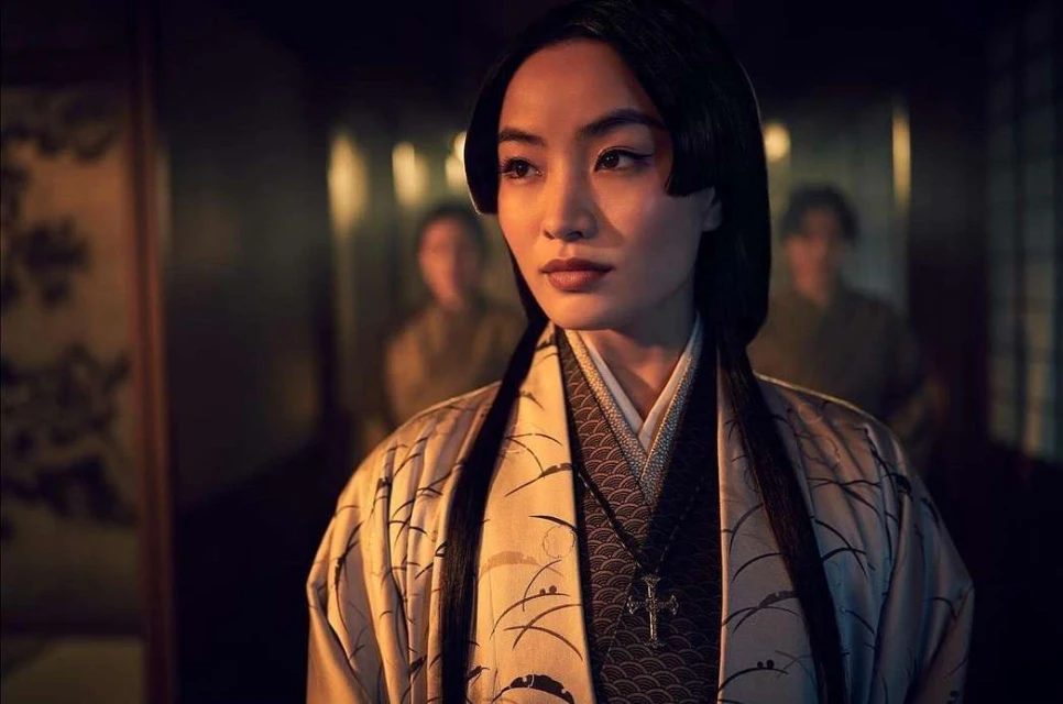 Portrait of an Asian woman with dark hair wearing a patterned kimono and a cross necklace indoors with people in the background.