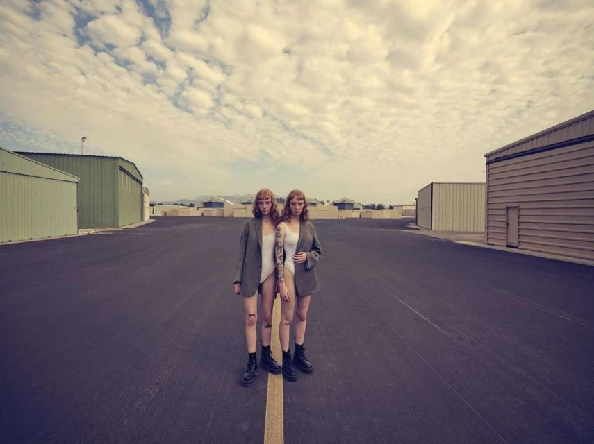 Two women in blazers and bodysuits stand on a tarmac runway against a backdrop of buildings and a cloudy sky.