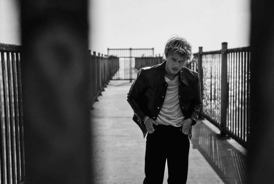 Black and white portrait of a stylish young adult male posing on a boardwalk or pier with a leather jacket and hands in his pockets.