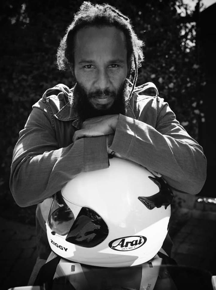 Monochrome portrait of a bearded man with dreadlocks in a jacket holding a motorcycle helmet in front of a blurred background.