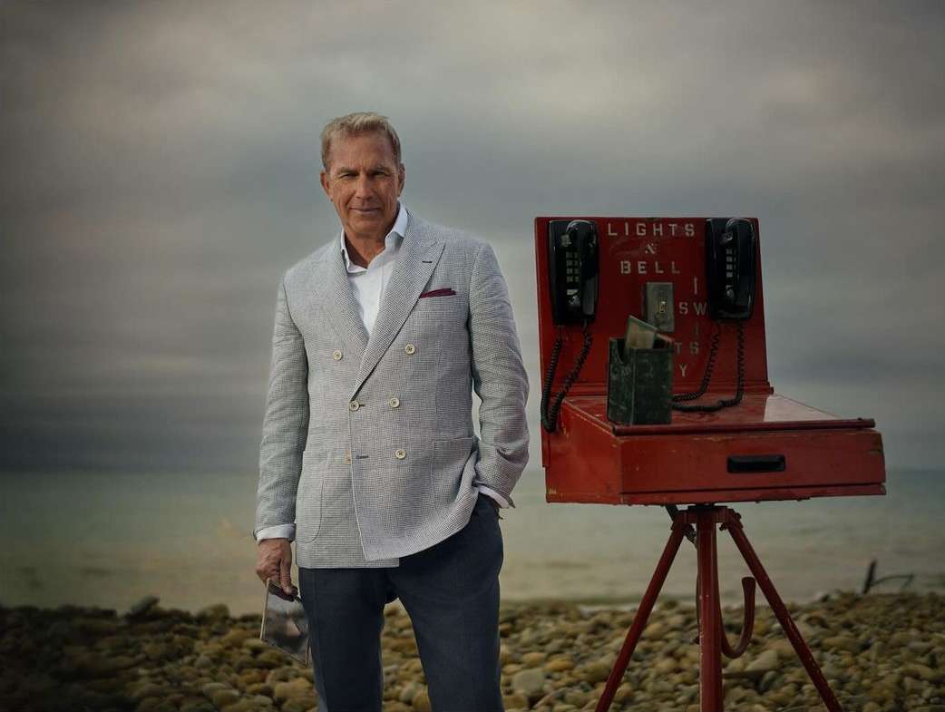 Adult male in a blazer stands near a red utility box with telephones on a rocky beach, showcasing fashion against a coastal backdrop.