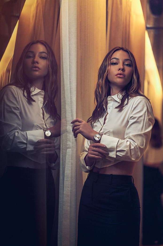 Attractive young woman in a white blouse and dark skirt poses indoors by a window, looking confidently at the camera with neutral expression.