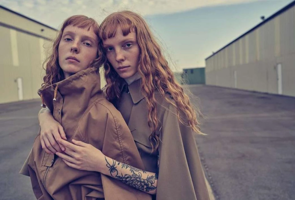 Two red-haired female models pose in matching brown jackets against an industrial building backdrop on an overcast day.