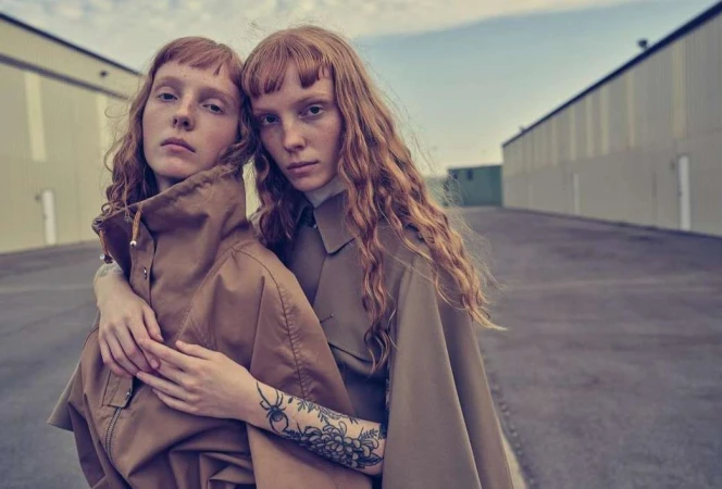 Two red-haired female models pose in matching brown jackets against an industrial building backdrop on an overcast day.