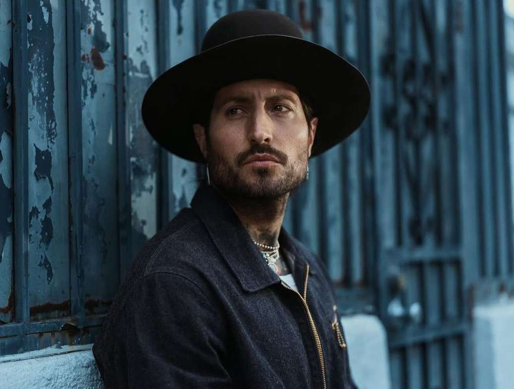 Portrait of a bearded adult male in a black hat and denim jacket posing against a blue textured wall, looking off to the side.