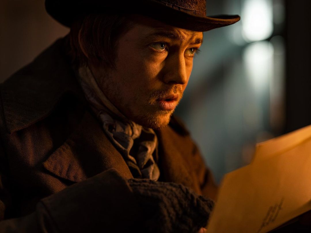 Close up of a man in a hat and coat reading documents, set indoors with warm lighting and a slightly concerned facial expression.