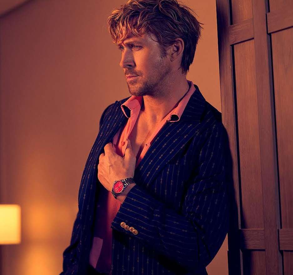 Profile shot of a fair-skinned adult man wearing a pinstripe jacket and pink shirt, leaning against a wood panel, looking left.