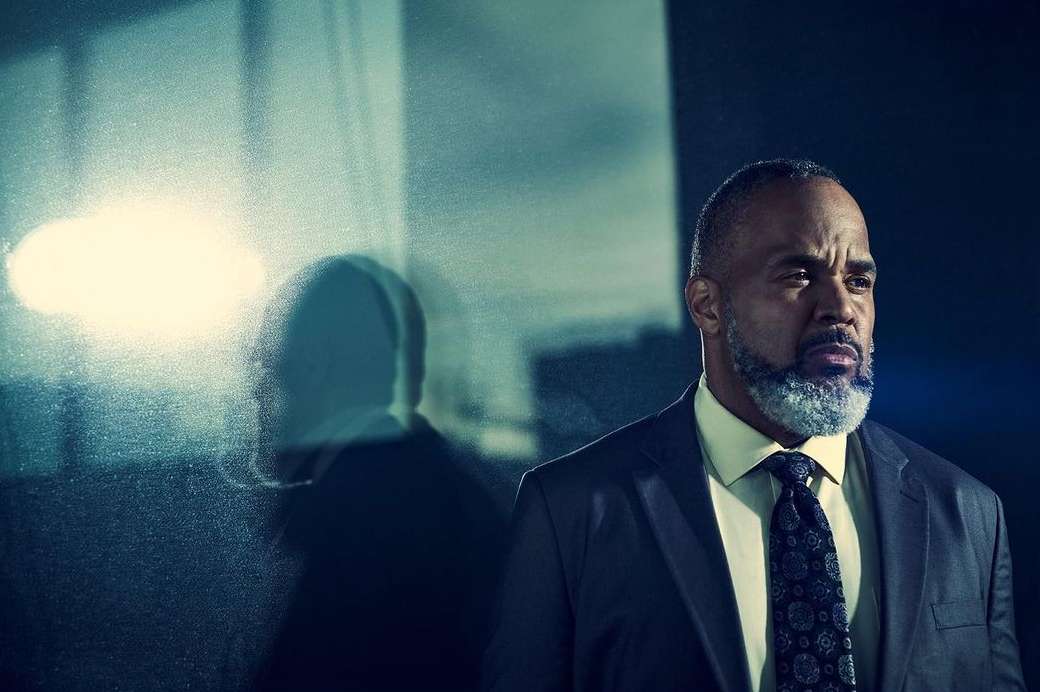 Portrait of a mature Black man in a dark suit with a patterned tie standing beside a glass panel reflecting his silhouette.