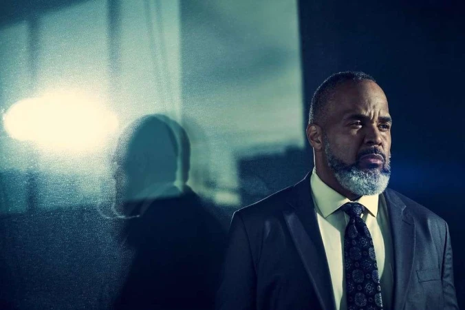Portrait of a mature Black man in a dark suit with a patterned tie standing beside a glass panel reflecting his silhouette.