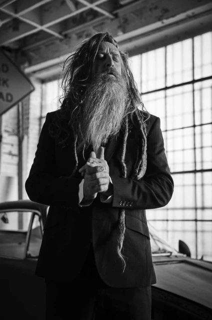 Monochrome portrait of an adult male with a long beard and dreadlocks wearing a suit in an industrial-style interior.