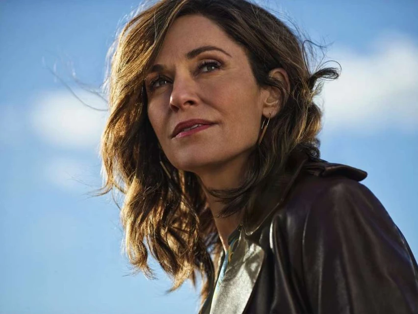 Portrait of a confident adult woman with brown hair, wearing a leather jacket, set against a bright blue sky with soft clouds.