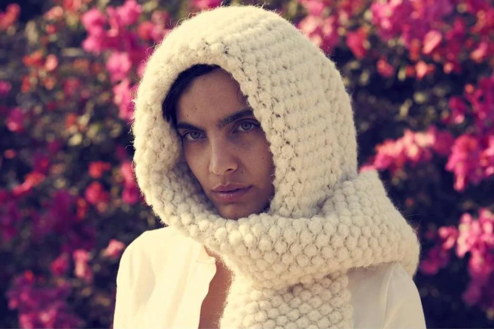 Portrait of a young adult female wearing a white knit hood and shirt, against a backdrop of pink flowers, creating a soft, natural look.