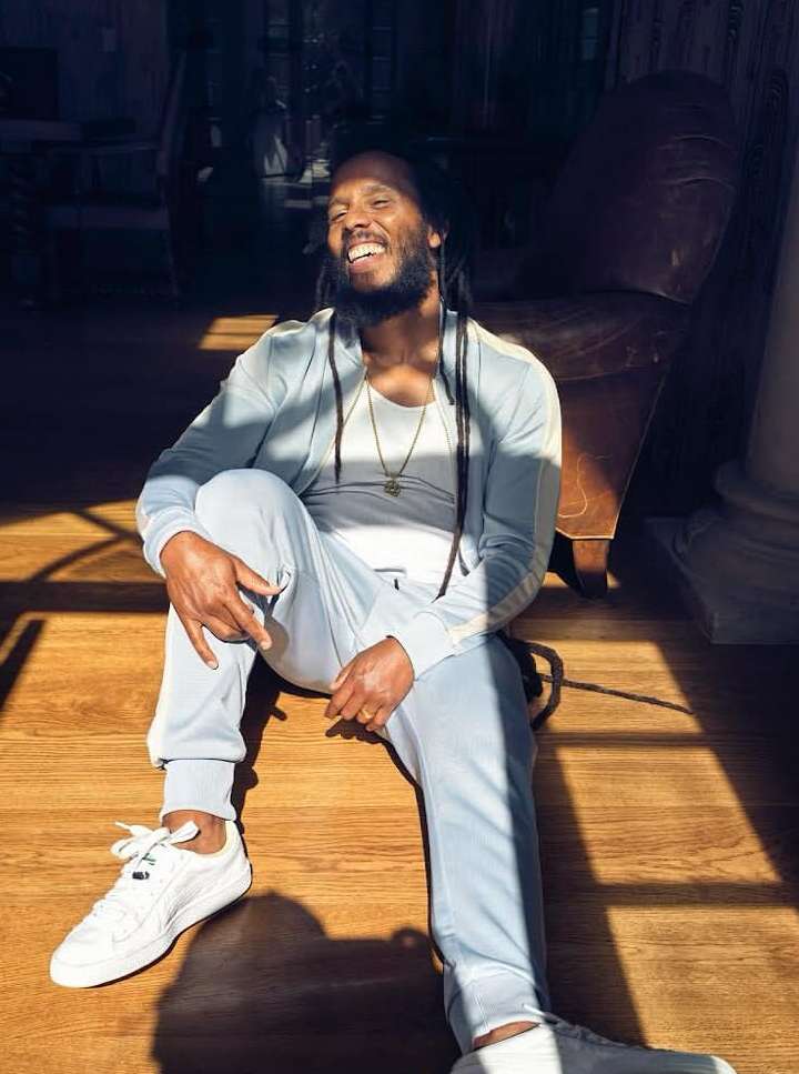 Smiling adult male with dreadlocks wearing a tracksuit and sneakers sitting on the floor in an indoor setting with natural light.