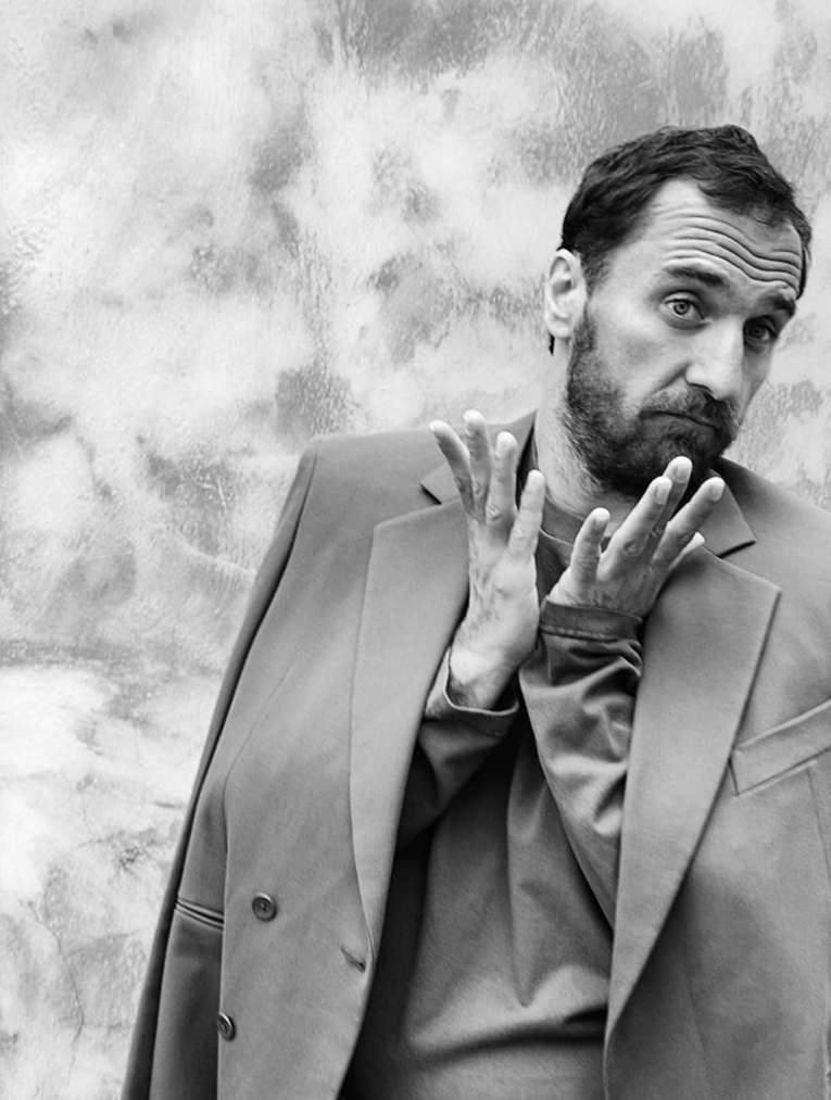 Black and white portrait of a man in a suit jacket, standing against a textured wall, looking at the camera with his hands raised.