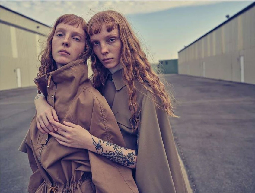Two women with auburn hair stand closely together outdoors in matching brown outfits against an industrial building backdrop.