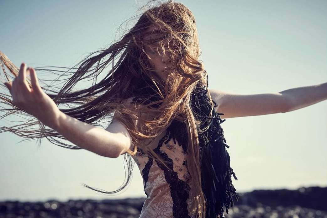 Woman with long brown hair wearing an ornate vest with outstretched arms on a rocky beach with a clear blue sky in the background.