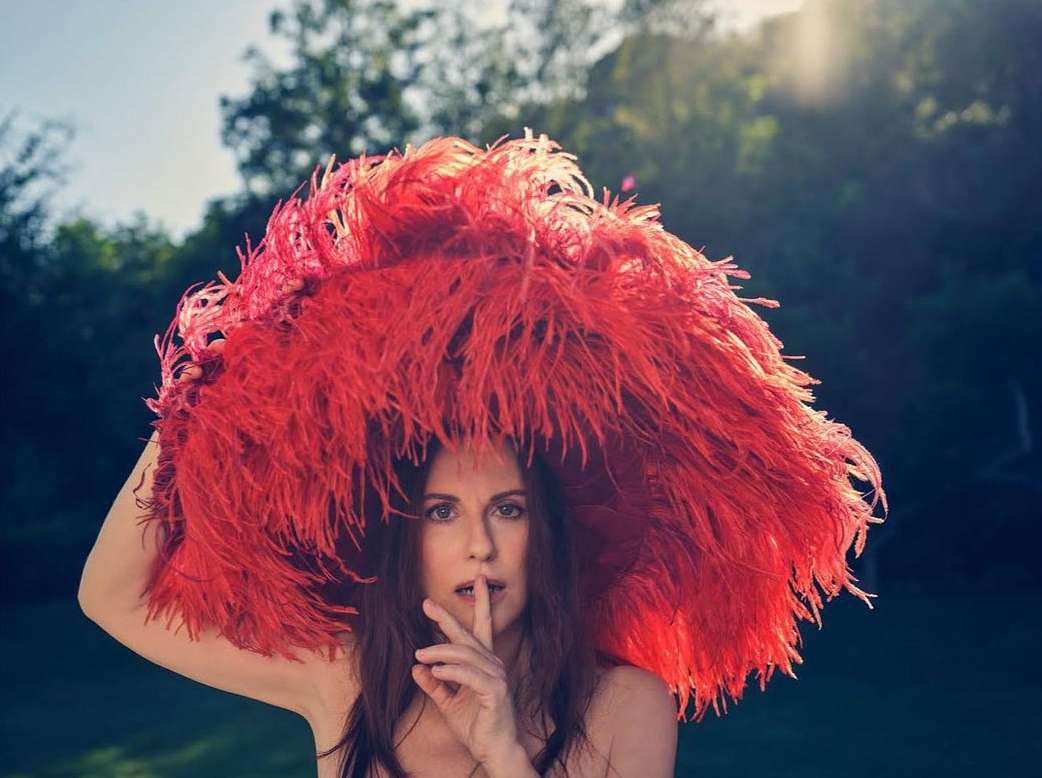 Adult female wearing a large, bright red feather hat, holding a finger to her lips in a shushing gesture, standing outdoors.
