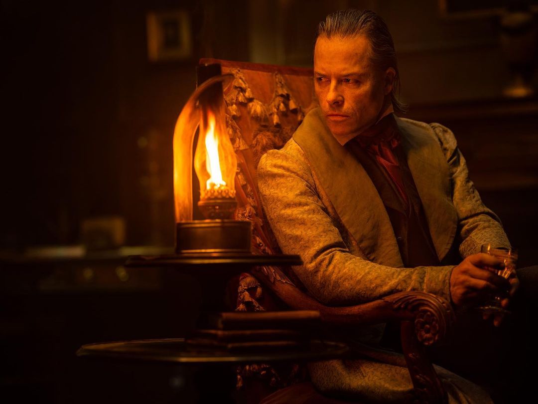 Dramatic portrait of an adult male in vintage attire holding glass next to lit oil lamp in dark, moody indoor setting.