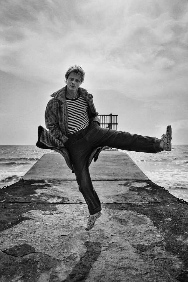 Monochrome portrait of a person jumping mid-air on a pier, wearing a striped shirt, coat, and shoes, with the ocean and sky in the background.