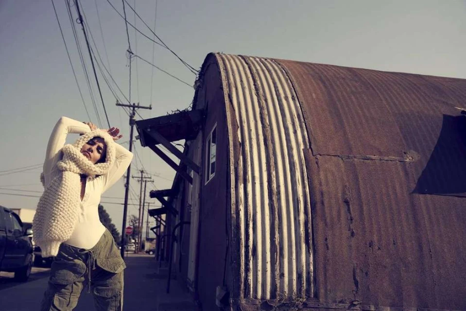 Woman poses with a scarf in an urban alleyway, featuring weathered metal buildings and overhead wires against a muted sky.