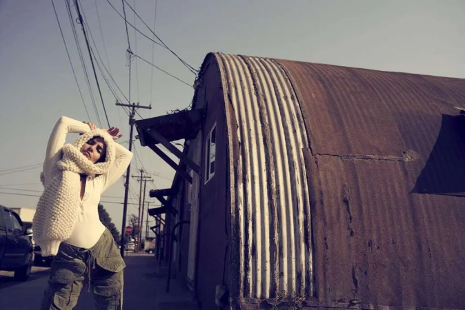 Woman poses with a scarf in an urban alleyway, featuring weathered metal buildings and overhead wires against a muted sky.