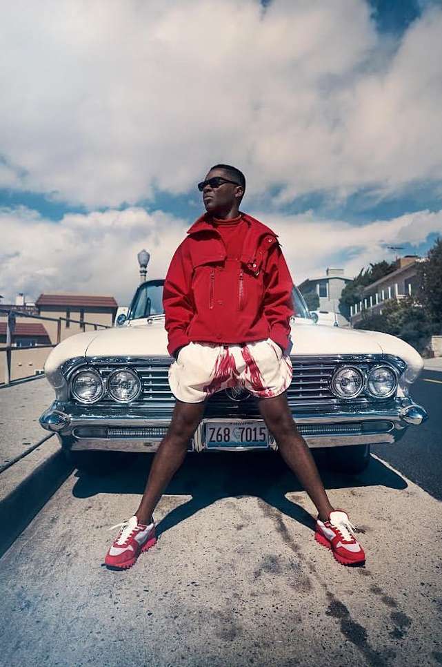 Adult male poses in front of a classic white car on an outdoor urban street with a cloudy sky, wearing sunglasses, a red jacket and red sneakers.