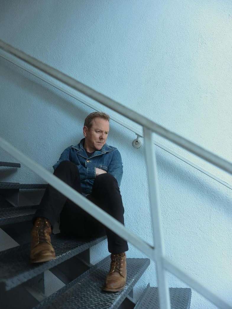 Man sitting on metal stairs with a denim jacket and brown boots. Blue wall in the background, creating a contemplative mood.
