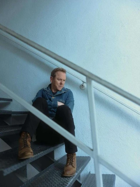 Man sitting on metal stairs with a denim jacket and brown boots. Blue wall in the background, creating a contemplative mood.