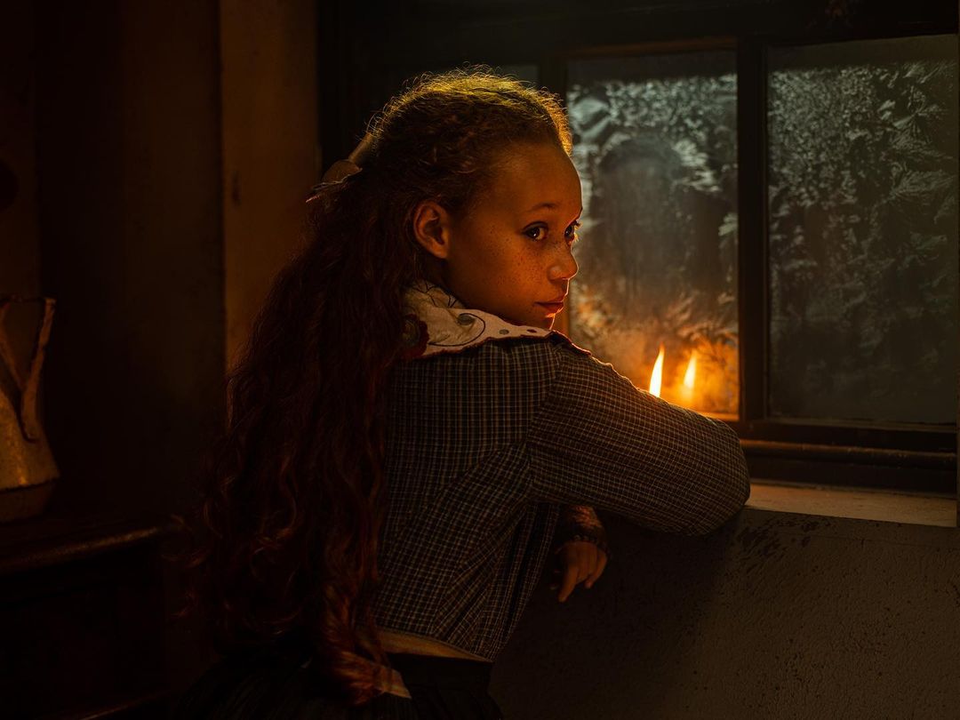 Young girl gazes out window in dim light. A candle sits on the windowsill, casting warm light on her face in the dark interior.