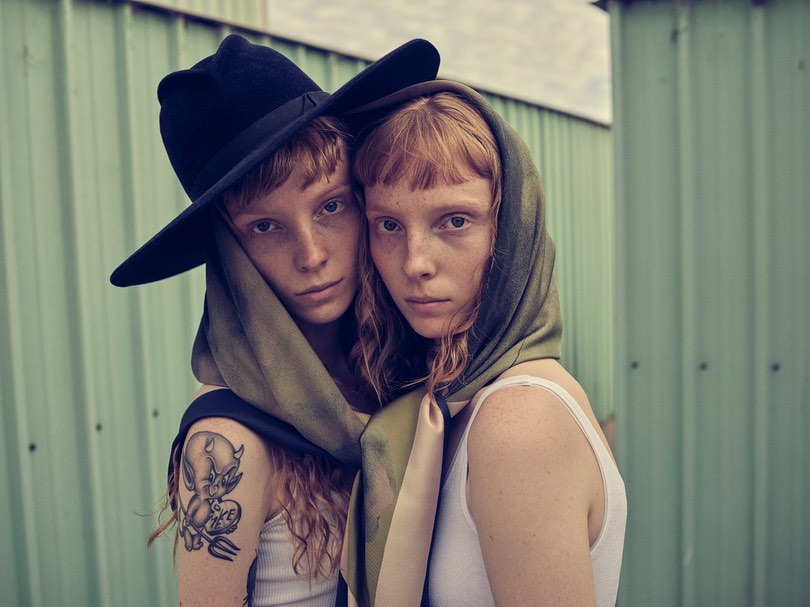 Two young women pose closely together against a corrugated metal background, one wearing a wide-brimmed hat and the other a headscarf.