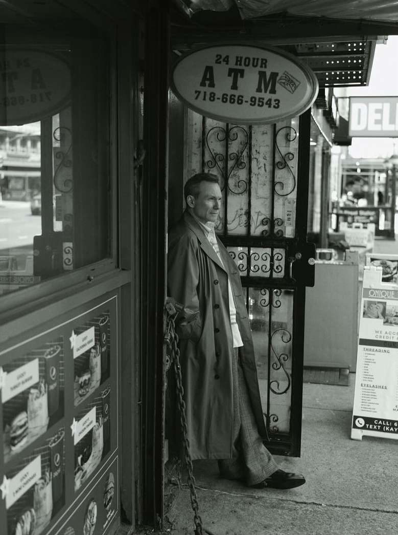 Black-and-white portrait of a mature man in a trench coat, standing in a doorway with an ATM sign, creating a candid, urban feel.