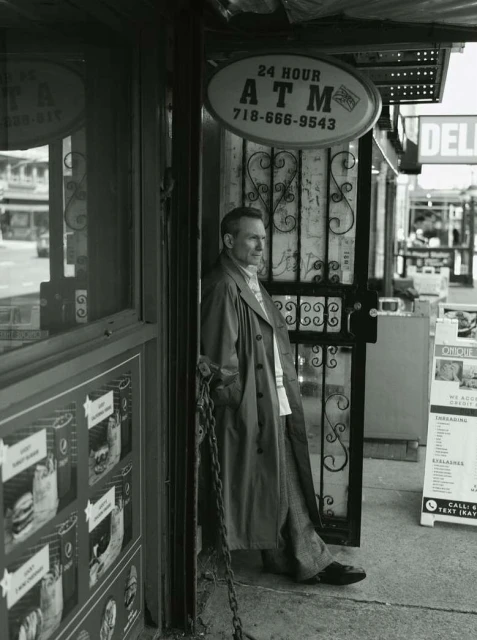 Black-and-white portrait of a mature man in a trench coat, standing in a doorway with an ATM sign, creating a candid, urban feel.