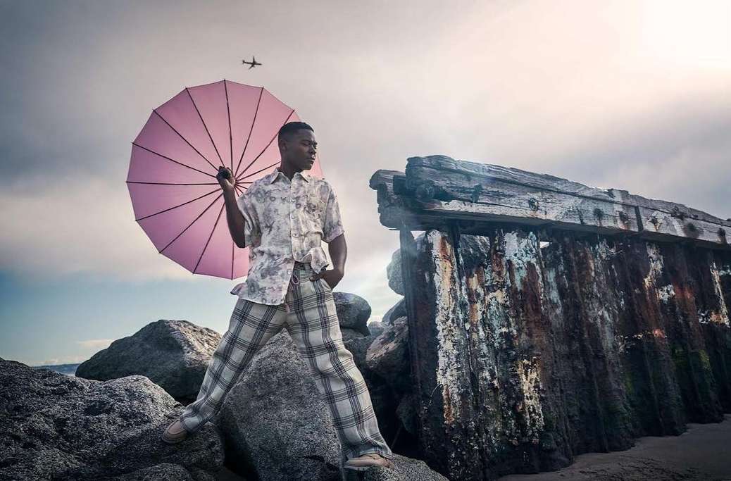 Man with umbrella stands on rocks near weathered wall, plane in sky. Stylish outfit, outdoor fashion photoshoot with neutral colors.