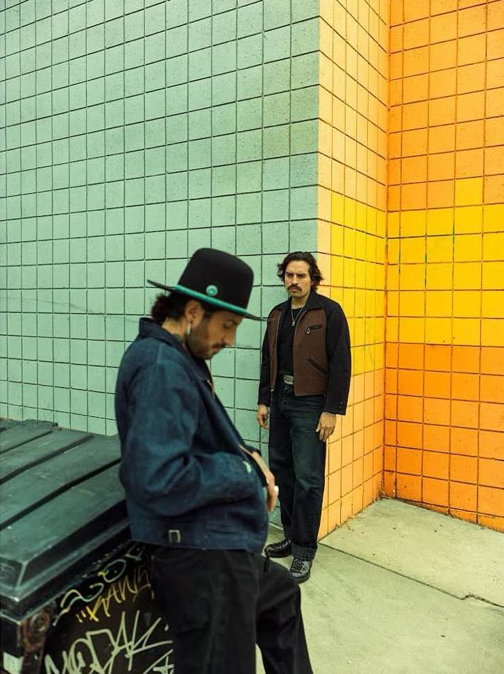Two men posing outdoors by a colorful tiled wall. One man in a hat leans on a container, while the other stands in the background.